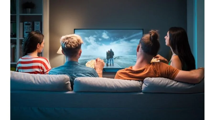 Four young adults watching television on a couch in a dimly lit living room.