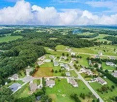 Aerial view of a rural area with houses, green fields, trees, and a large pond in the distance under a partly cloudy sky.