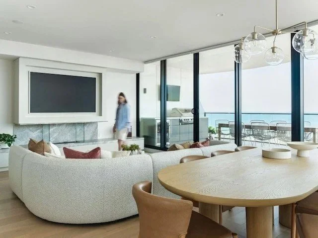 Modern living room with a large flat-screen TV, curved white sofa with cushions, dining table with chairs, and floor-to-ceiling windows with a balcony view.