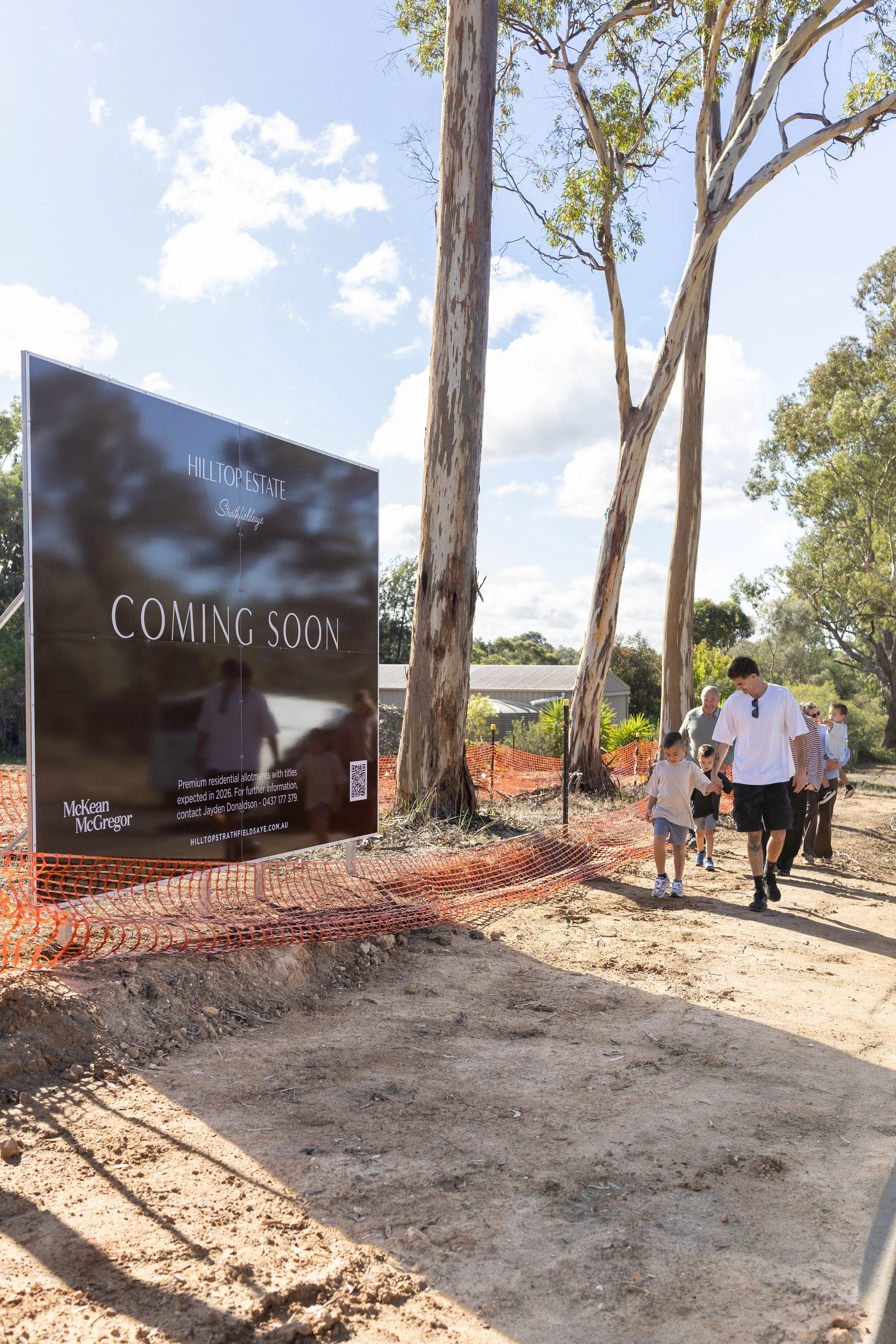 A group of people walking along a construction site with trees and a sign that reads "Hilltop Estate Coming Soon." The sky is partly cloudy, and there is orange construction fencing at the site.