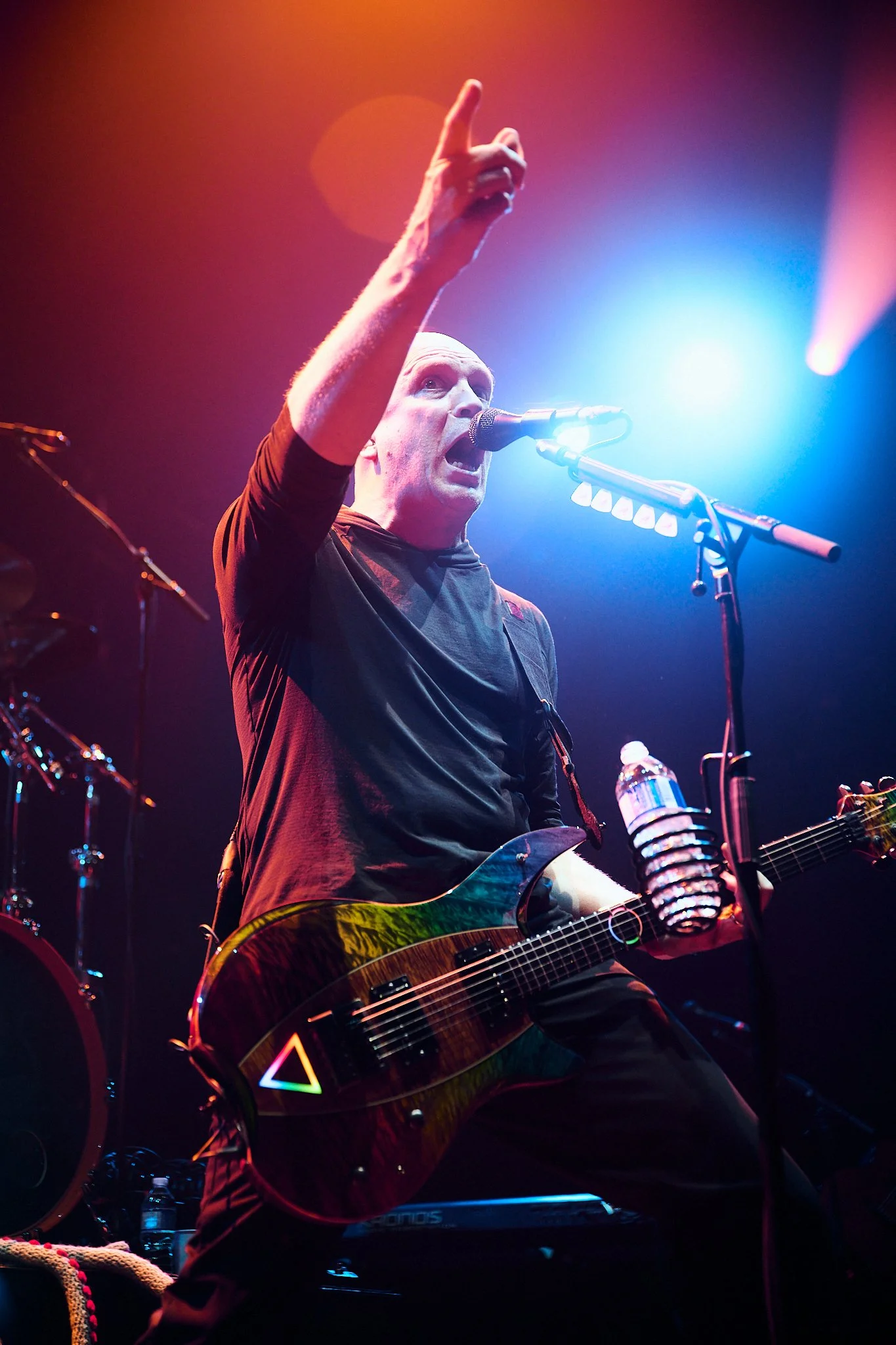 A male musician performs on stage with a guitar, singing into a microphone. He is wearing a black shirt, and is raising his right hand, gesturing upward. The stage is lit with colorful lights, including blue and orange.