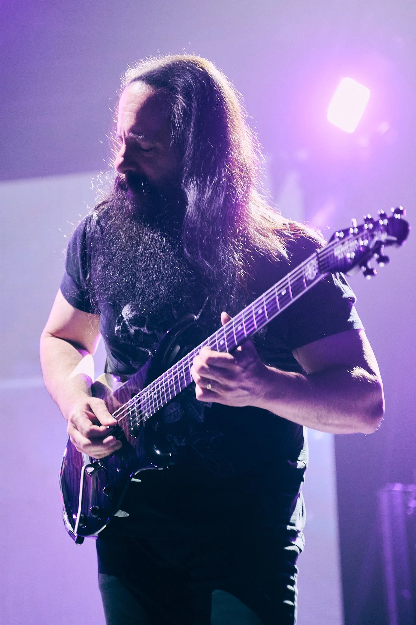 A man with long hair and a beard playing an electric guitar on stage, illuminated by purple stage lights.