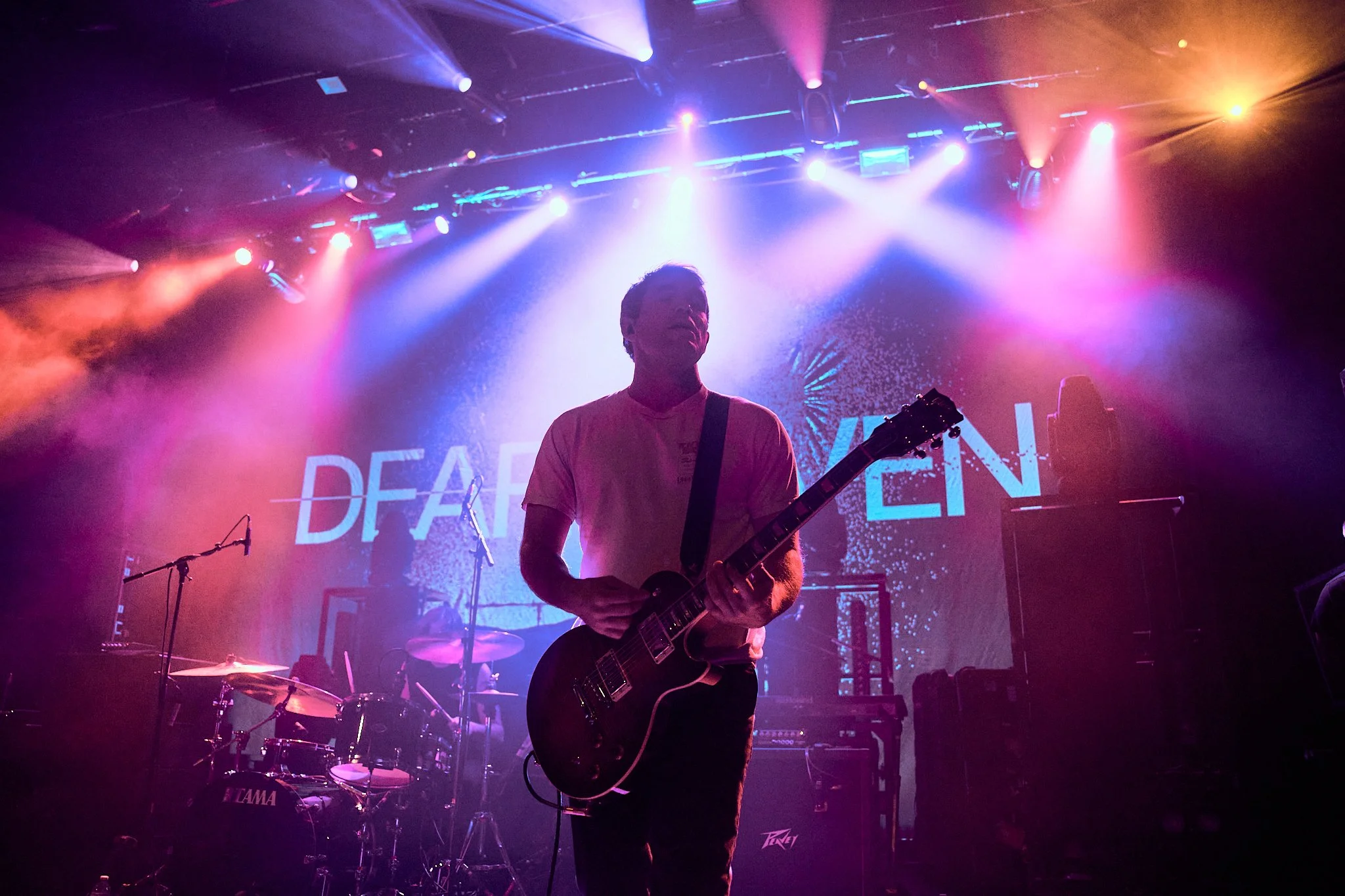 A musician playing an electric guitar on stage with colorful lights overhead and a screen in the background displaying the partially visible text 'DEAFEN'.