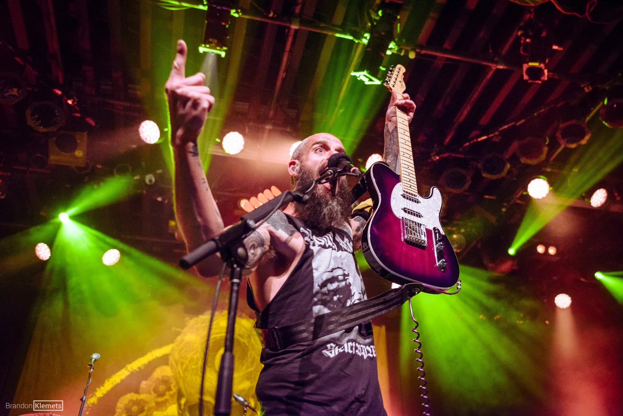 A man with a beard and tattoos on his arms is performing on stage with a pink electric guitar. He is raising his left hand with his index finger pointing upward while singing into a microphone. The stage has vibrant yellow and green lighting with a b