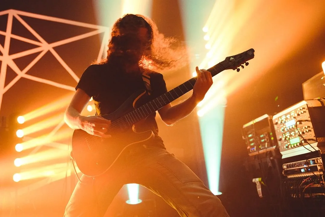 A person playing an electric guitar on stage with colorful lighting and sound equipment in the background.