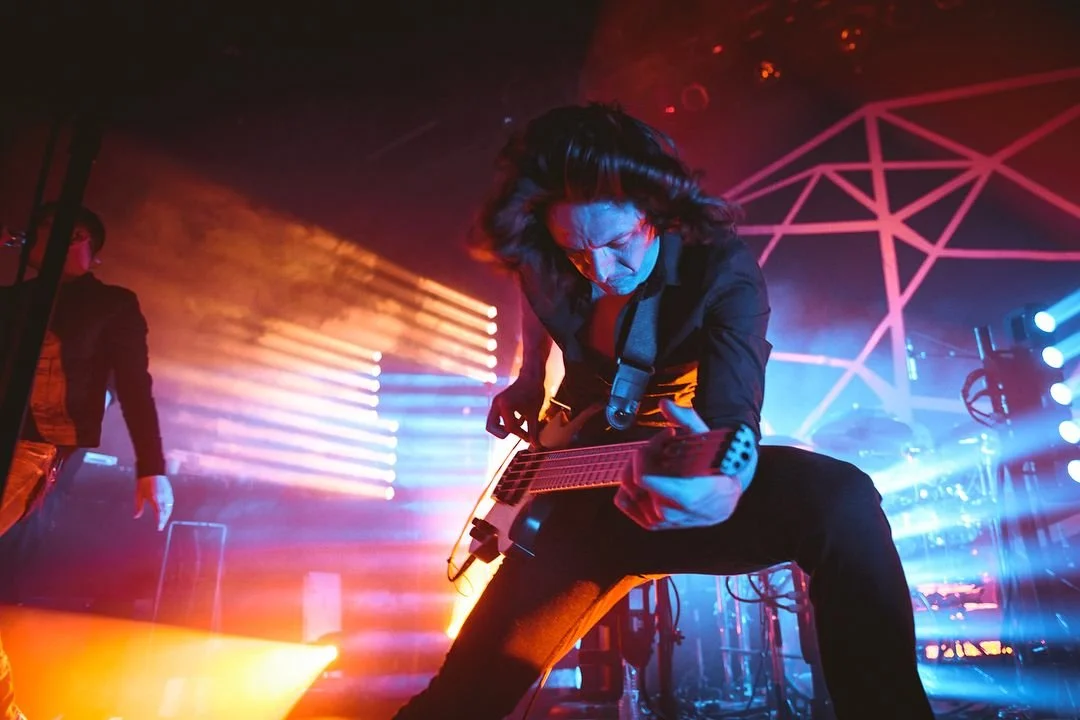 Musician playing electric guitar on stage with colorful lights and geometric background.