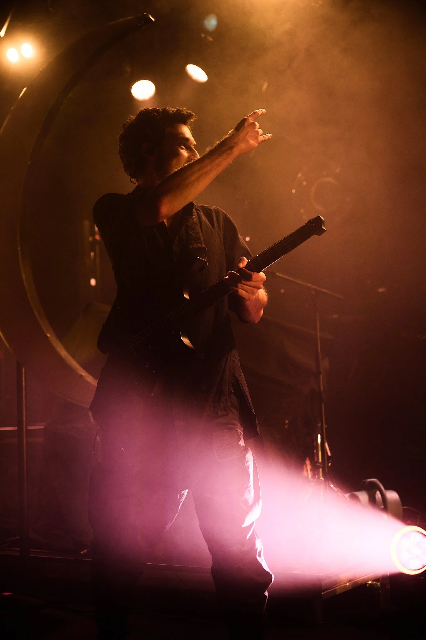 A man on stage playing electric guitar surrounded by orange and pink lights and smoke, with one hand raised and pointing.