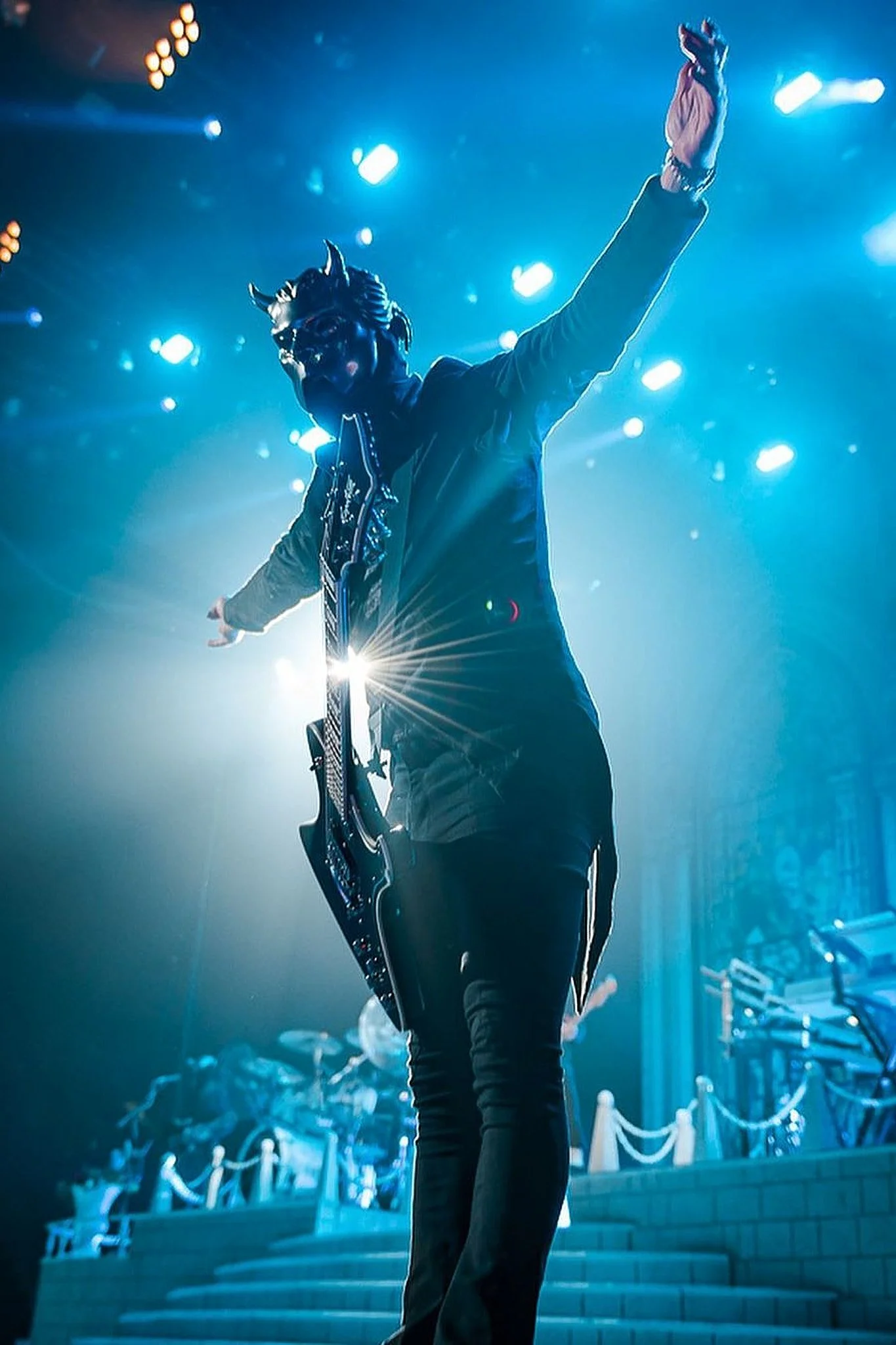 Person dressed in a devil mask and jacket, holding a guitar, on a stage with bright blue lights.