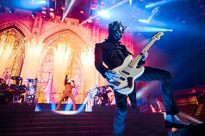 A masked musician playing a bass guitar on stage with a singer in the background, illuminated by colorful stage lights at a concert.