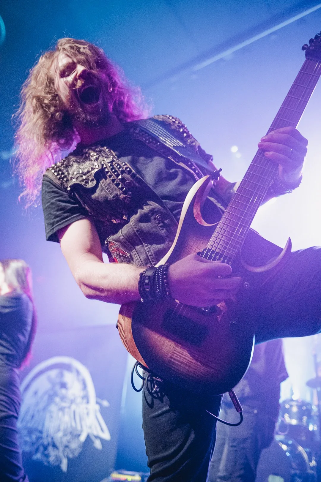A male guitarist with curly hair, wearing a black vest with spikes, passionately playing an electric guitar on stage with colorful stage lights.