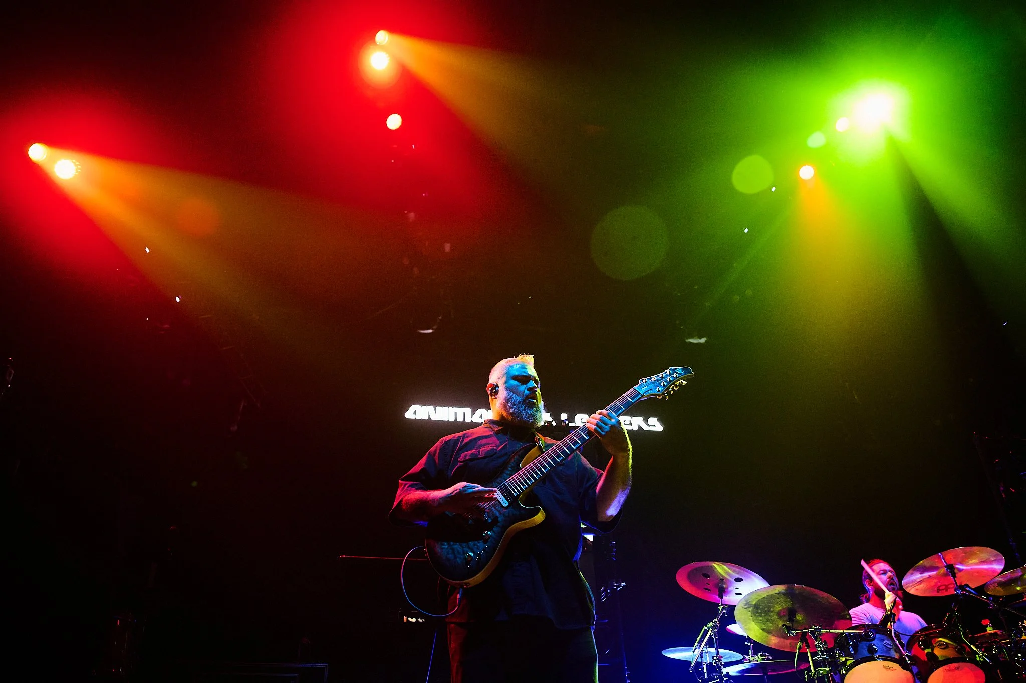 A guitarist and a drummer performing on stage under colorful red, yellow, and green lights.