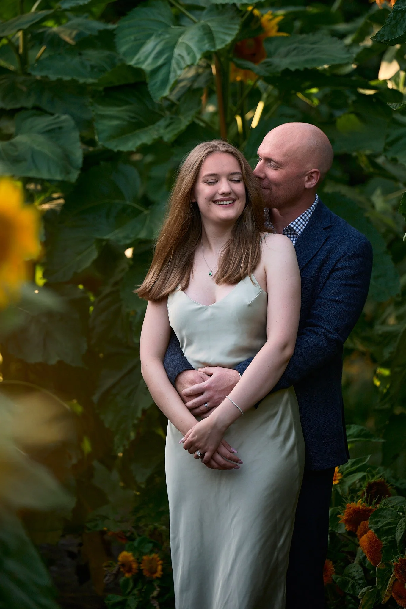A couple standing close together outdoors among large green leaves and yellow flowers, with the man embracing the woman from behind and both smiling.