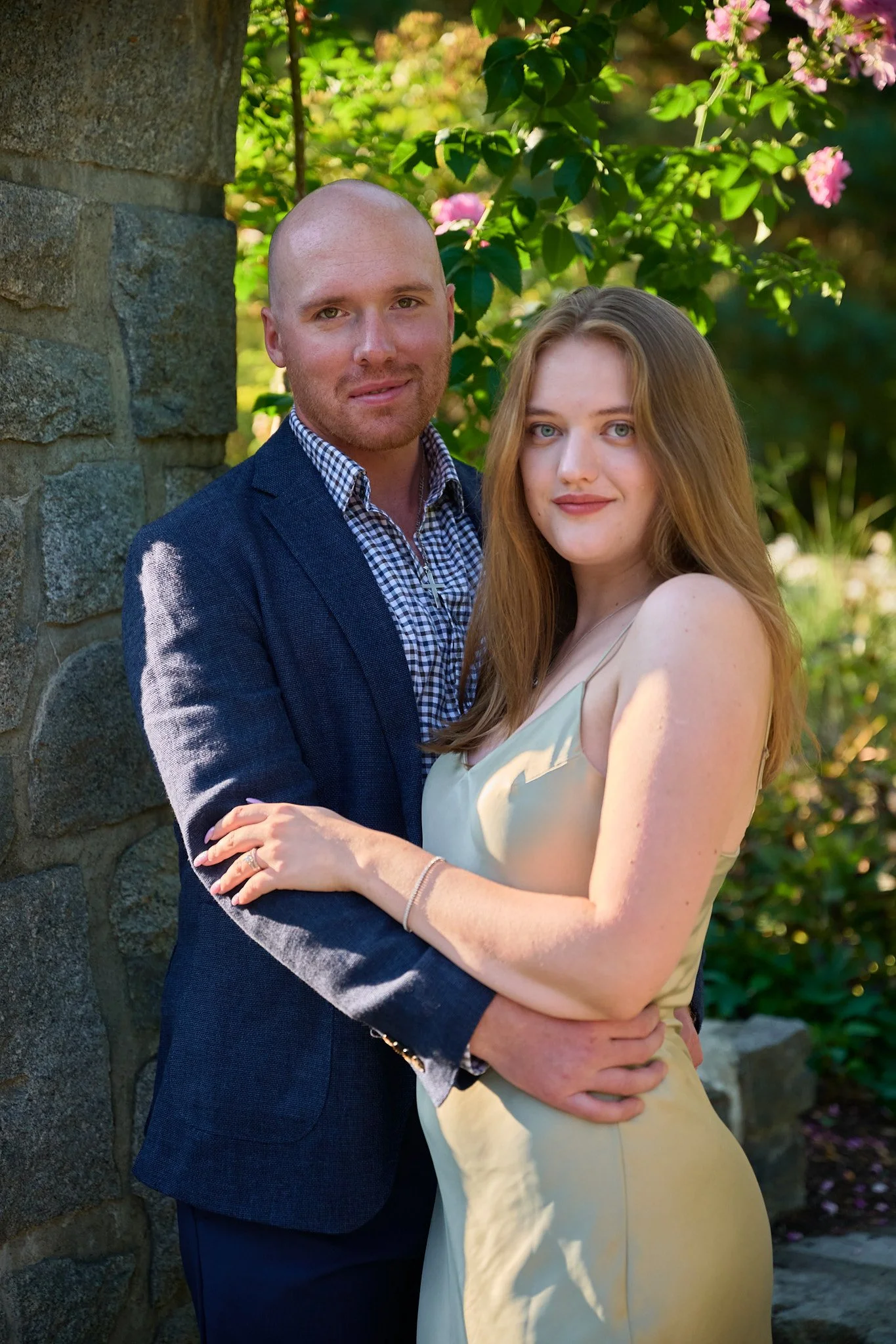 A couple posing outdoors, with the man wearing a dark blazer and checkered shirt, and the woman in a light-colored dress, standing close together with arms around each other, in front of green foliage and pink flowers.