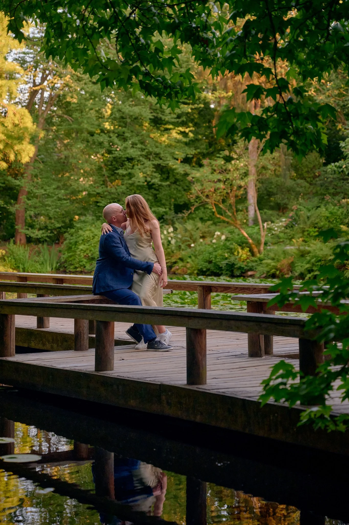 A couple sharing a kiss on a wooden bridge over a pond, surrounded by lush green trees in a park setting.