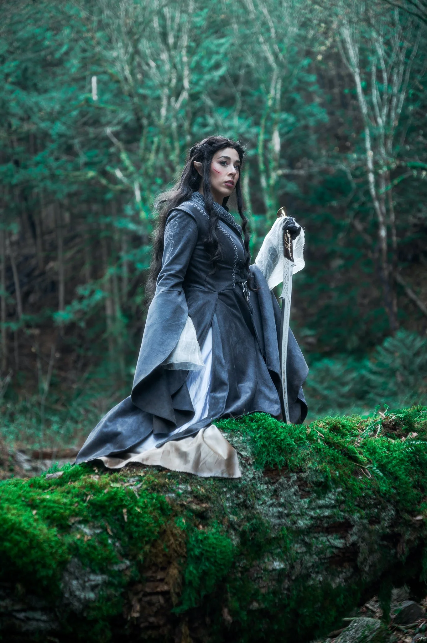 A woman dressed as an elf standing on a moss-covered log in a forest, holding a sword, with trees and foliage in the background.