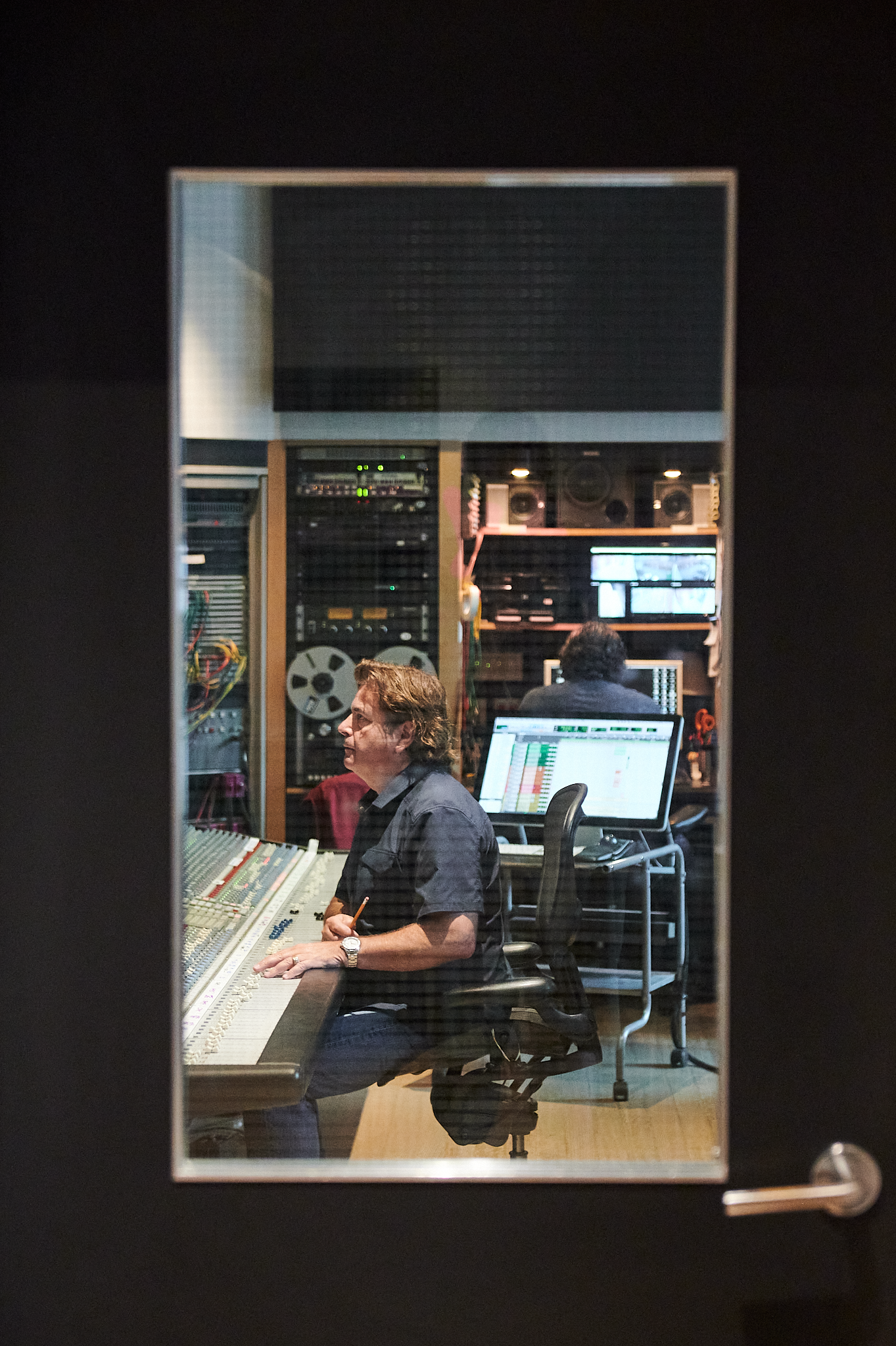 Studio engineer working at mixing console in recording studio, viewed through a glass door.