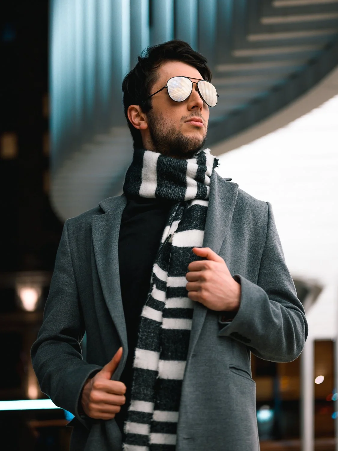 A stylish man wearing reflective sunglasses, a grey coat, and a black and white striped scarf, standing outdoors in an urban setting.
