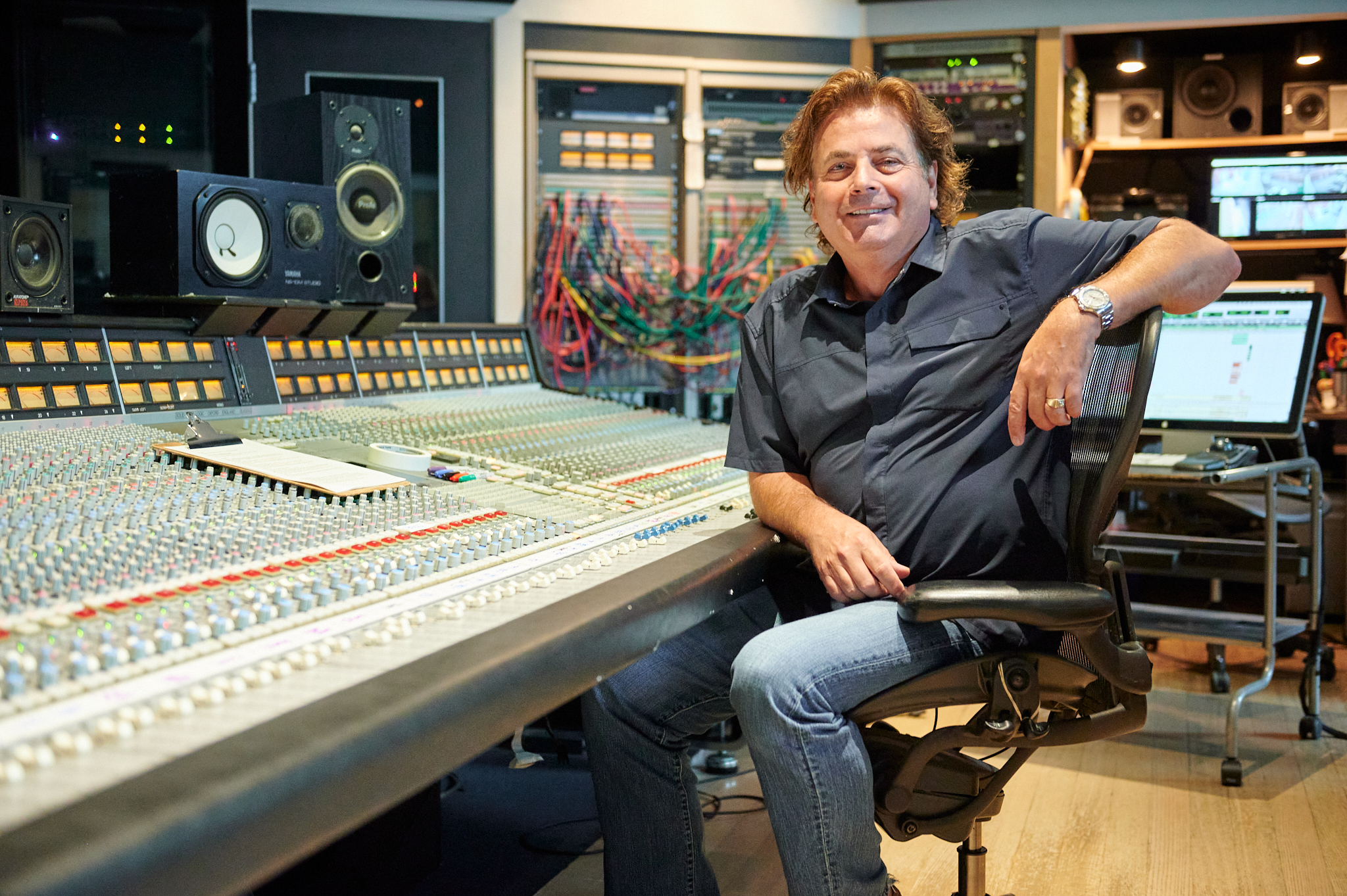 A man sitting in a recording studio next to a large mixing console with various equipment and monitors in the background.