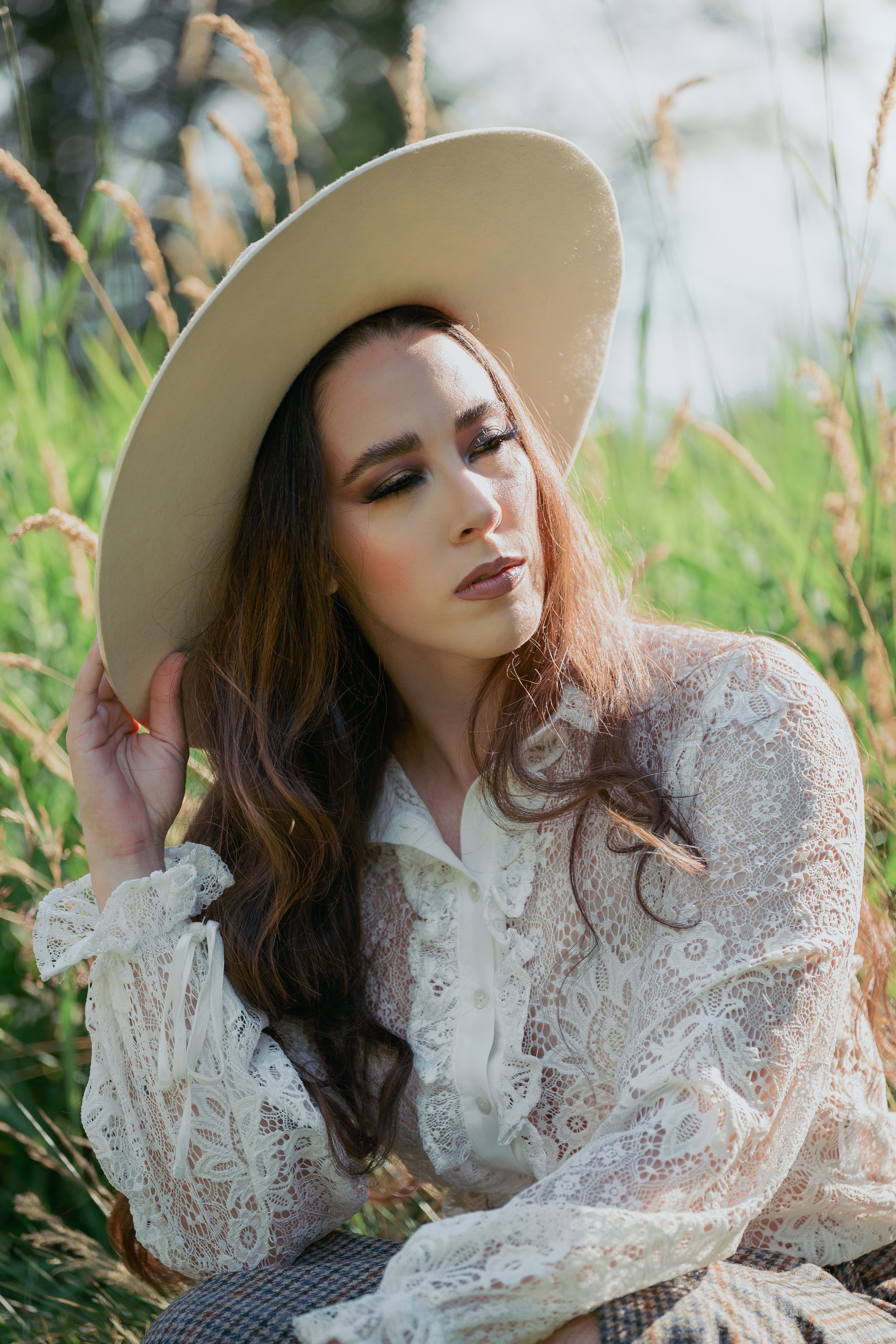 Woman with long wavy hair in a lace blouse and wide-brimmed hat sitting outdoors among tall grass, natural sunlight highlighting her face.