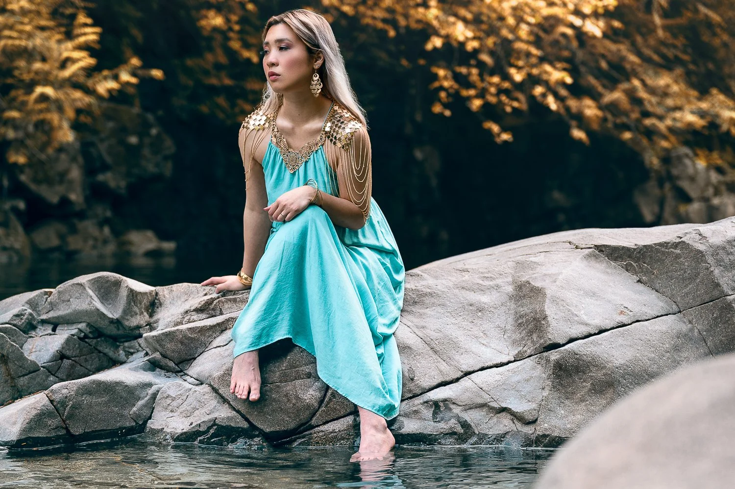 A woman in a flowing turquoise dress sitting on rocks beside a river, adorned with gold jewelry and a metallic shoulder piece, with autumn-colored foliage in the background.
