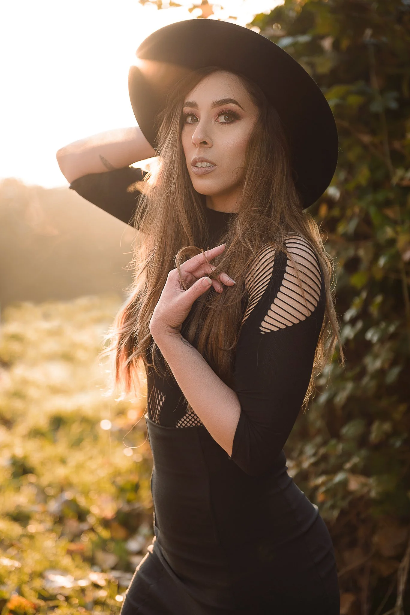 A woman with long brown hair wearing a black hat and a black dress with cutout details, standing outdoors during sunset.