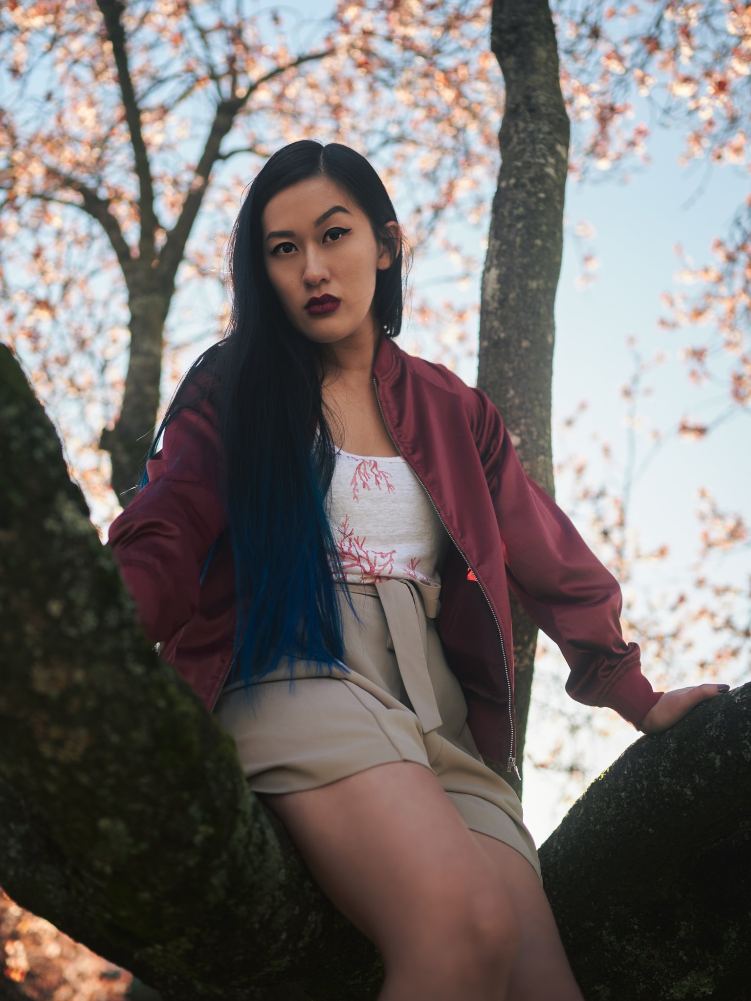 A woman with long black and blue hair sitting on a tree branch outdoors with autumn leaves and sky in the background.