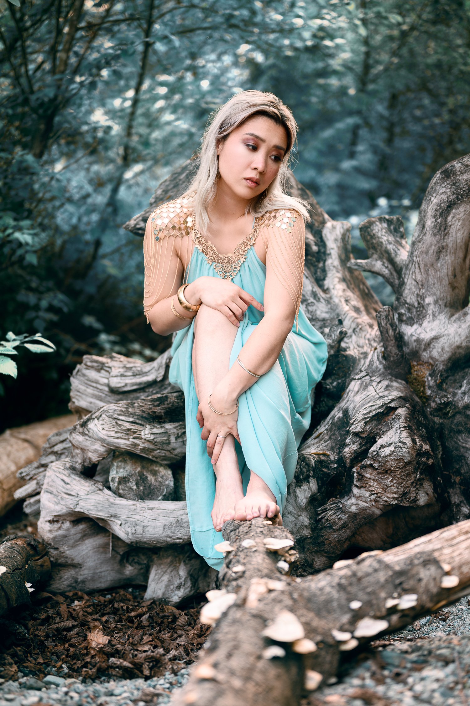 A woman with blonde hair sitting on a large fallen tree in a forest, wearing a flowing light blue dress and gold jewelry, looking contemplative.