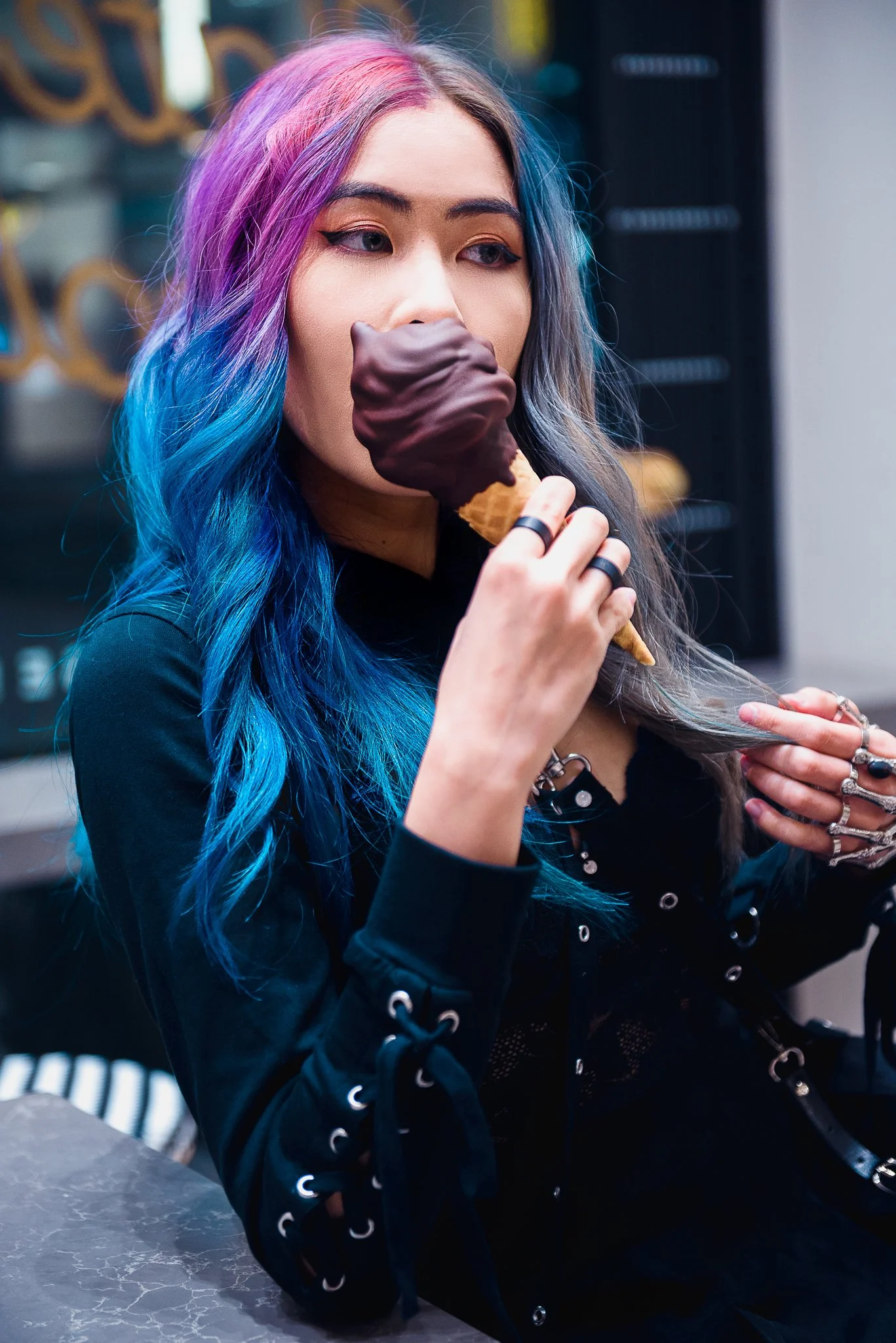 Young woman with colorful blue, purple, and pink hair, wearing black clothing, enjoying an ice cream cone with a chocolate-coated scoop.