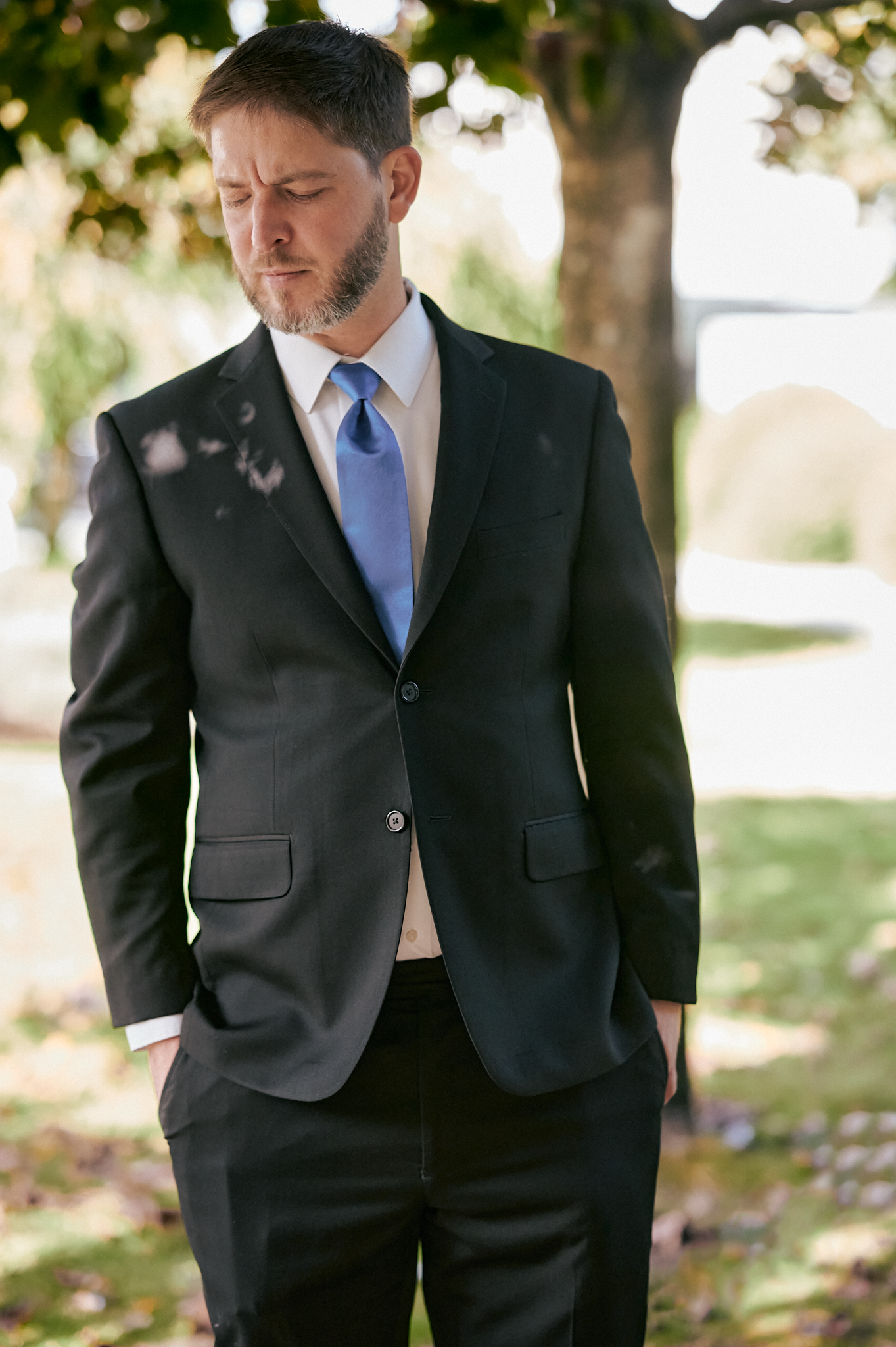 A man in a black suit with a blue tie, looking down with a contemplative expression, standing outdoors near a tree.