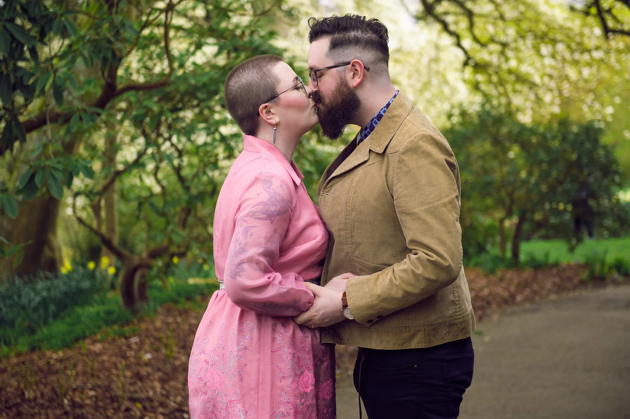 A couple sharing a kiss outdoors in a park with green trees and a dirt path.