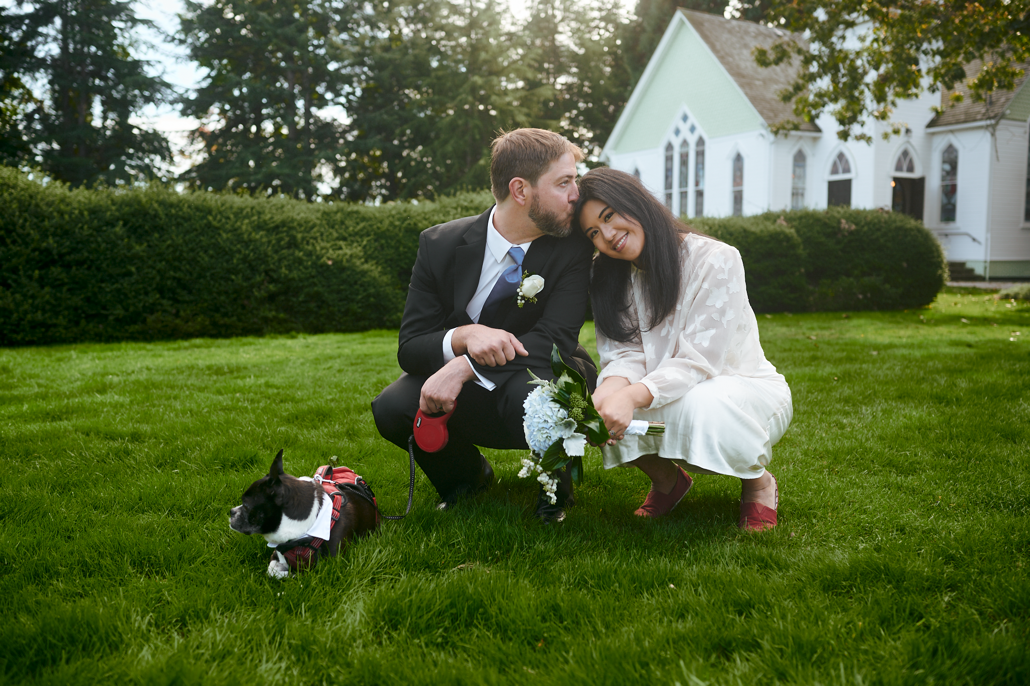 Couple, dressed in a tuxedo and white dress, sitting on the grass with a dog dressed in a bow tie, holding a bouquet of flowers, in front of a white church with green roof, surrounded by trees.