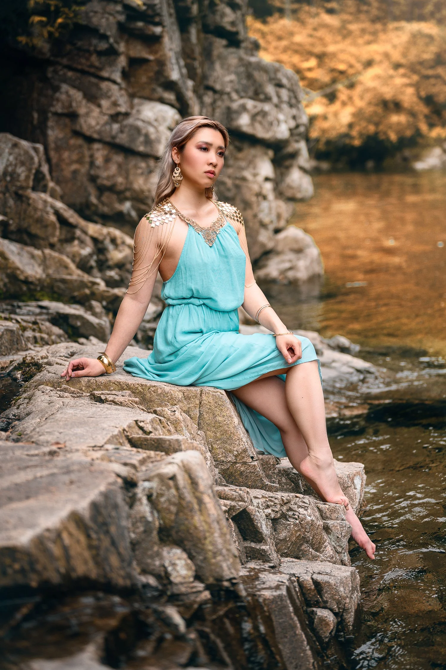 A woman in a turquoise dress sitting on rocks by a river with fall-colored trees in the background.