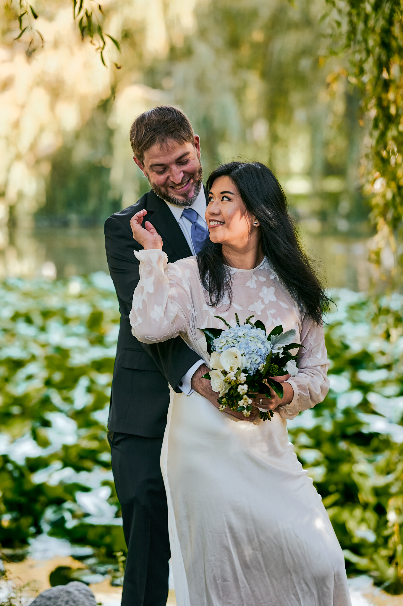 A bride and groom smiling and embracing outdoors near a pond with lily pads, trees, and sunlight filtering through the foliage.