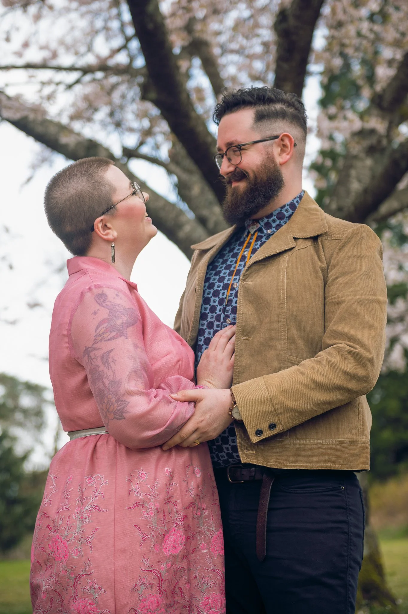 A couple standing closely together outdoors, gazing into each other's eyes, with trees and a cloudy sky in the background.