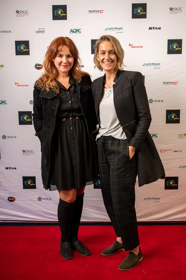 Two women standing on a red carpet in front of a step and repeat banner with various logos, smiling at the camera.