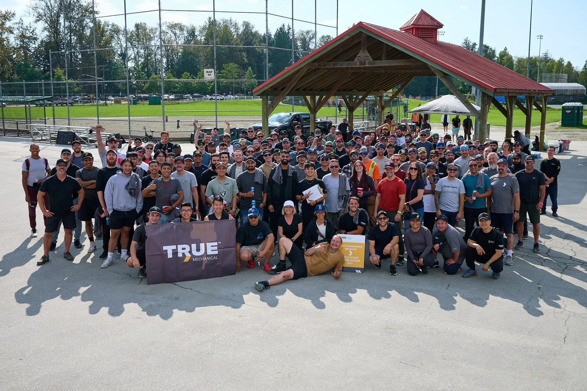A large group of people posing for a group photo outdoors near a baseball field, with some people holding banners and drinks.
