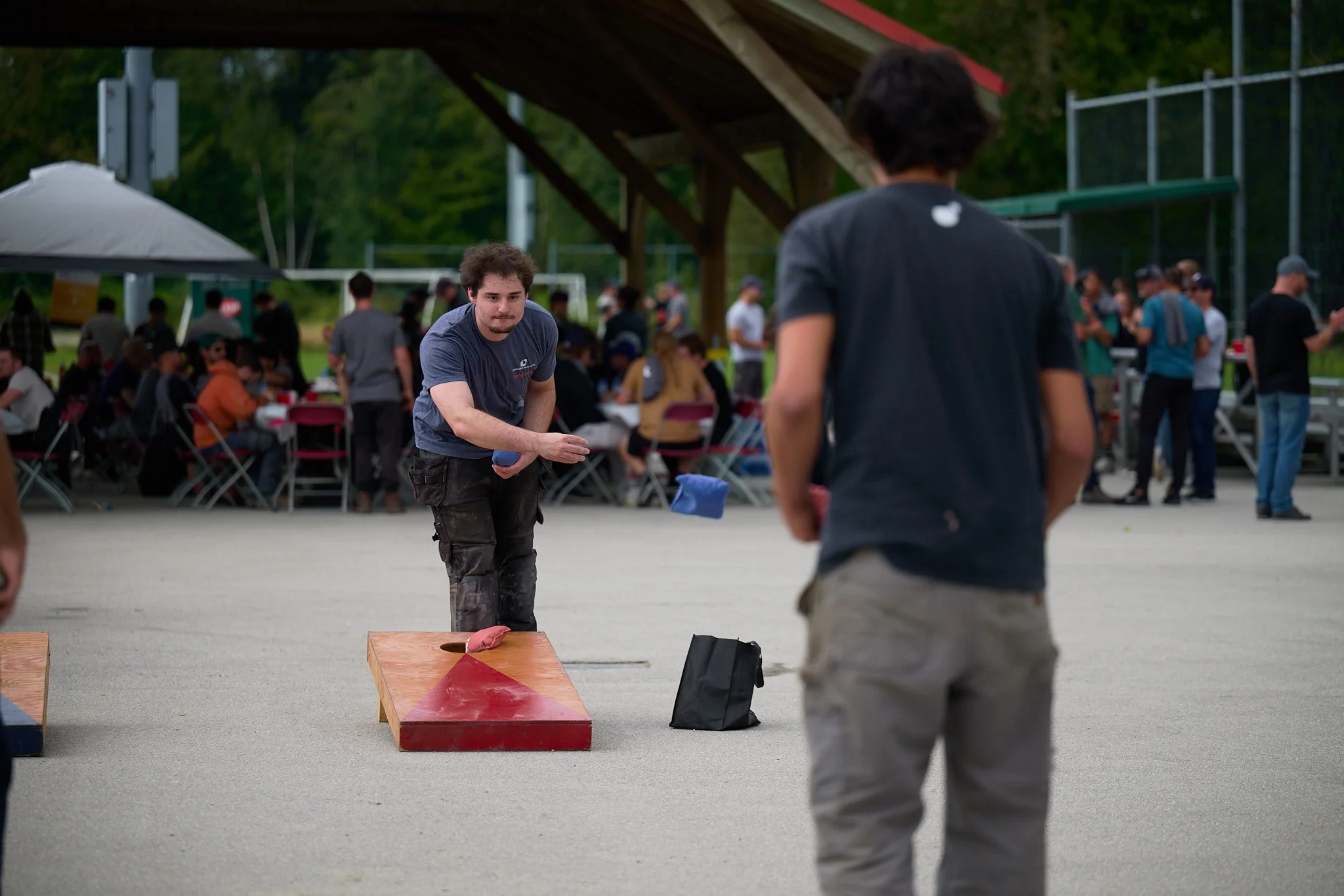 Two young men playing a game that involves throwing bean bags onto a wooden platform with holes, outdoors at a gathering with people sitting at tables and standing under a canopy in the background.