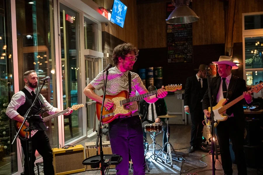 A band performing live in a cozy indoor venue with four members playing guitars and drums, with warm lighting and large windows showing outside lights.