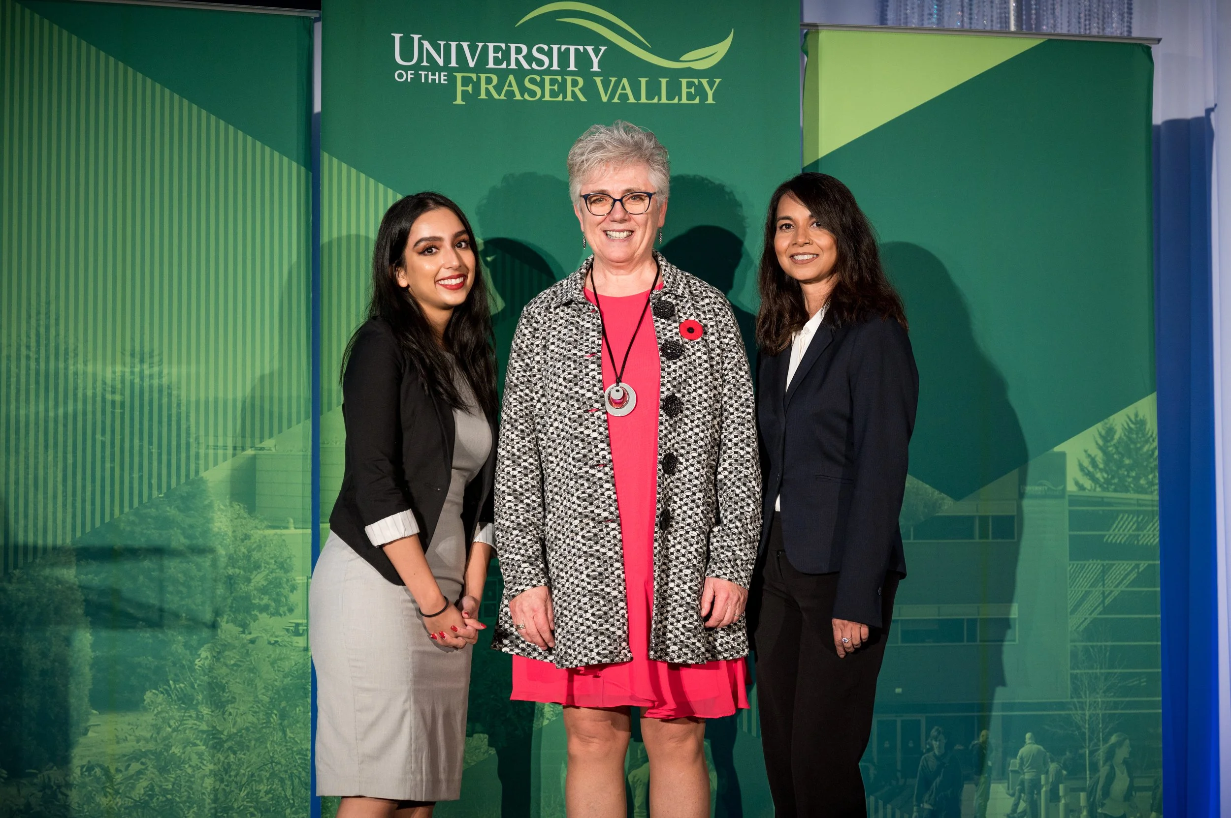 Three women posing for a photo at the University of the Fraser Valley, standing in front of a green backdrop with the university's name and logo. The woman in the middle is wearing glasses, a patterned coat over a pink dress, and a poppy pin. The wom