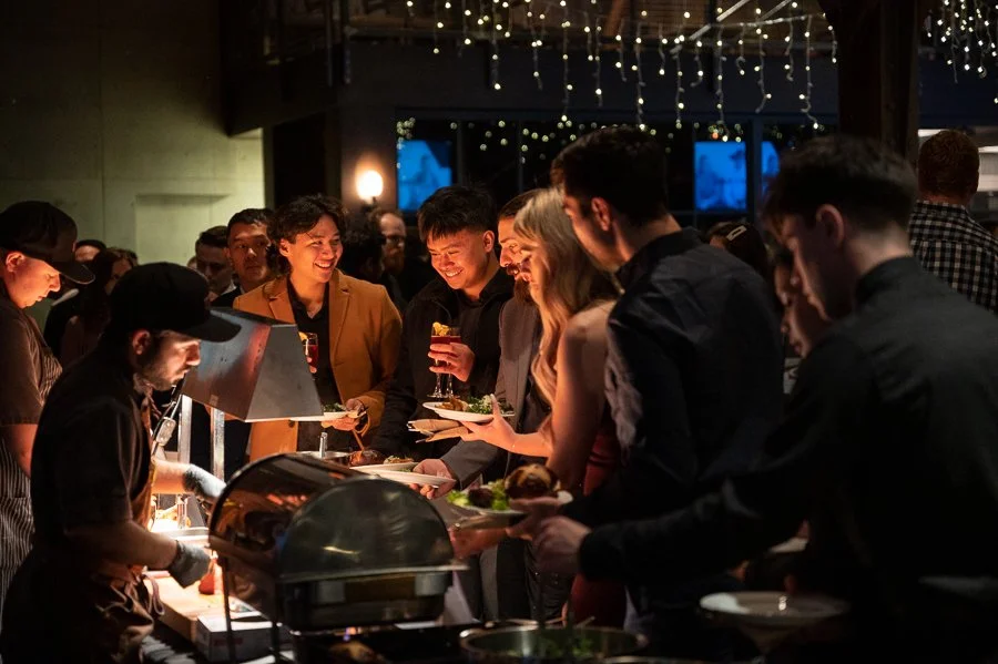 People at a buffet line enjoying food and drinks in a dimly lit venue decorated with string lights.