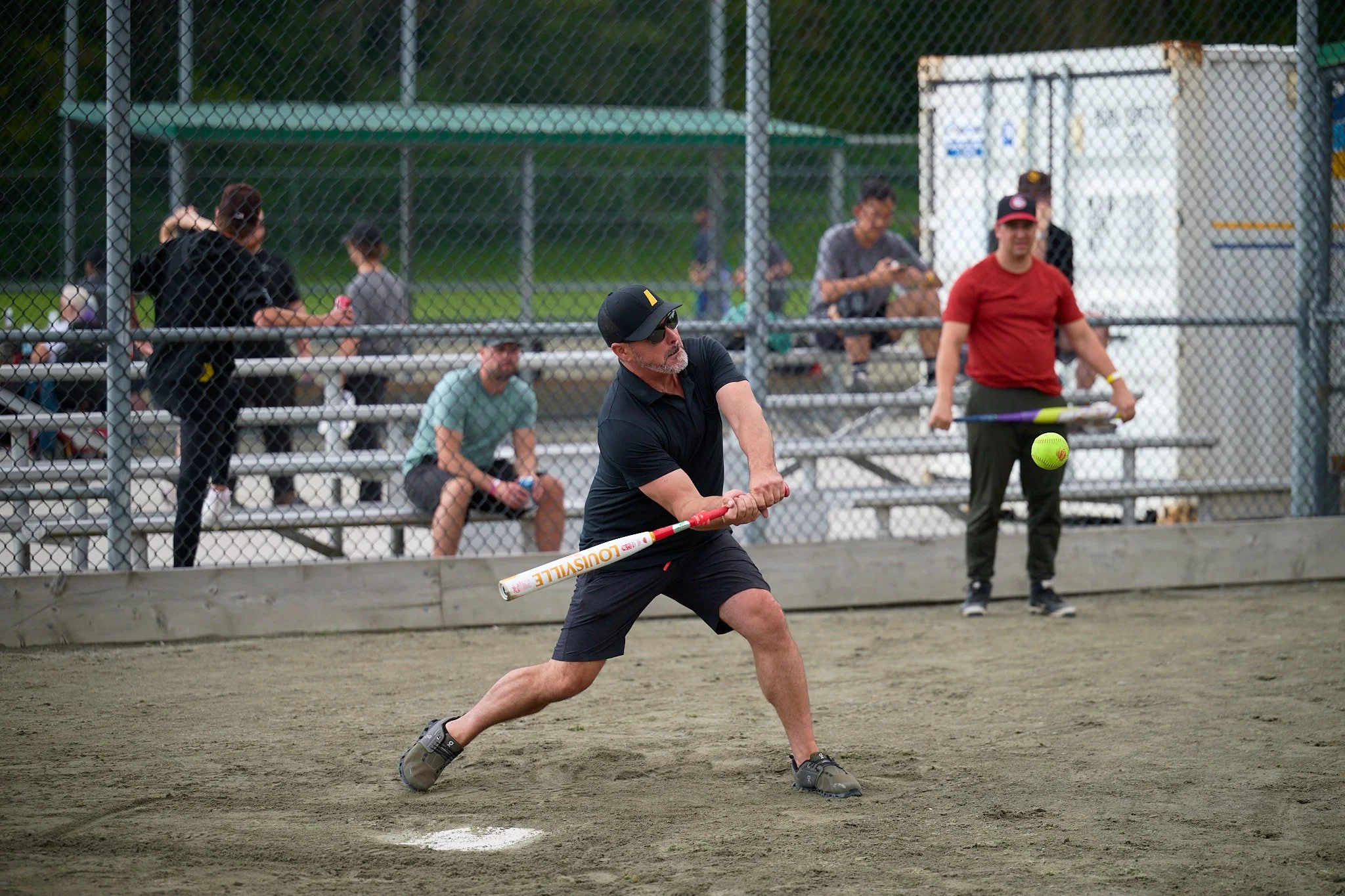 A man wearing a black cap, sunglasses, a black t-shirt, and shorts is swinging a red and white plastic bat at a green softball on a dirt field during a game, with spectators watching behind a chain-link fence.