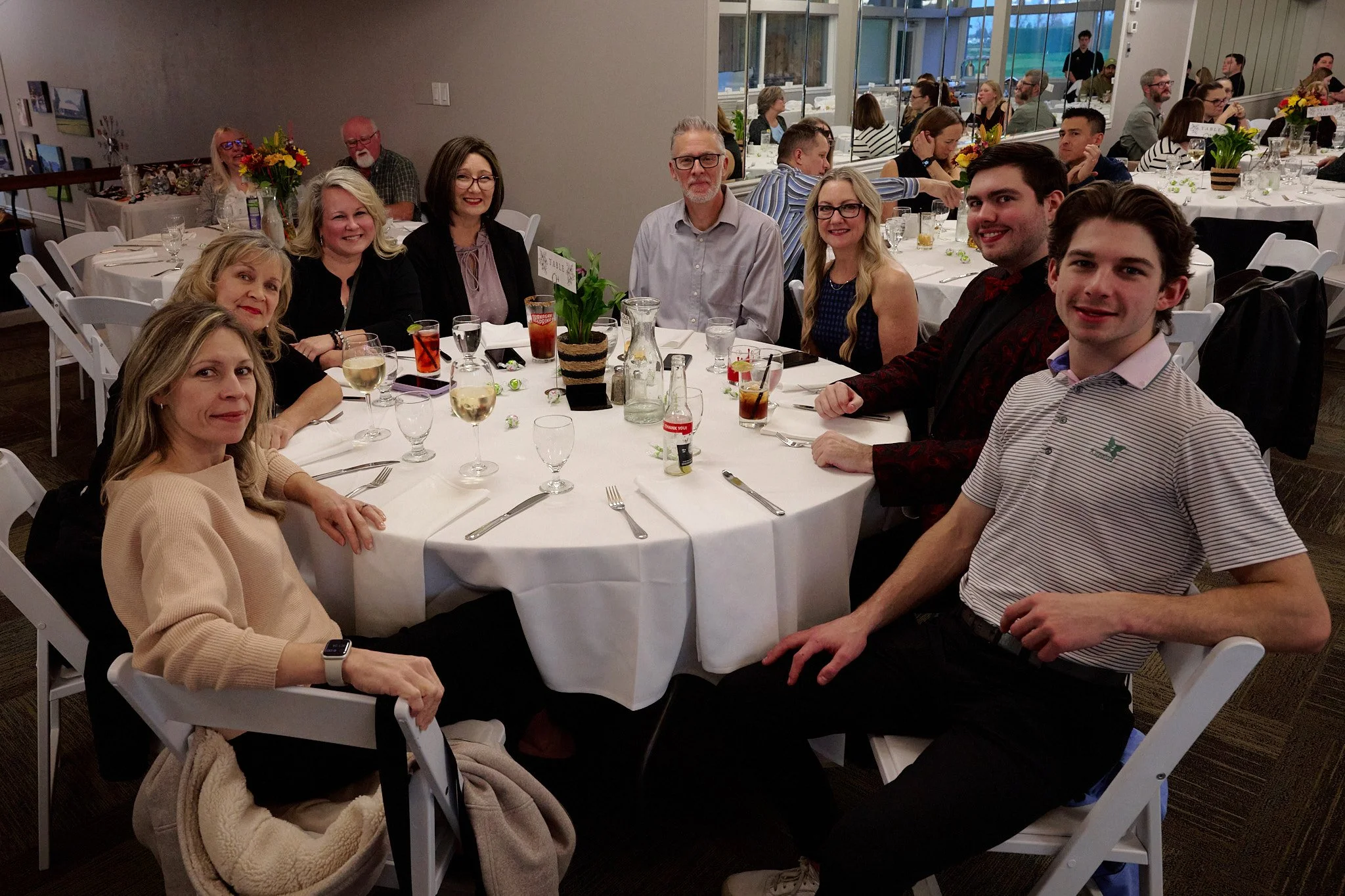 Group of eleven people seated around a dinner table at a formal event in a banquet hall, with glasses of wine and soft drinks, and floral centerpieces.