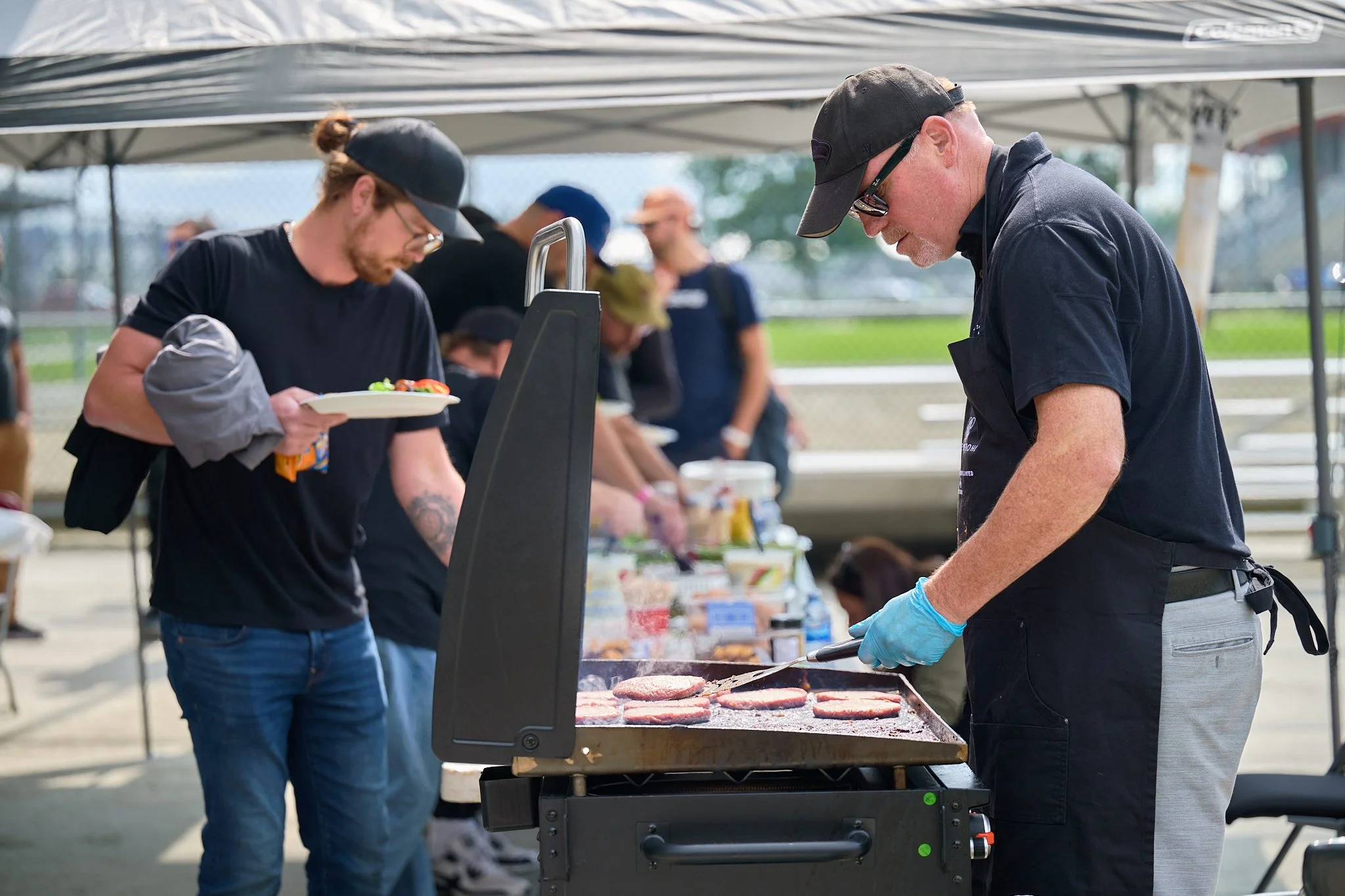 Chef grilling burger patties at outdoor barbecue, with people in line and a table of food in the background