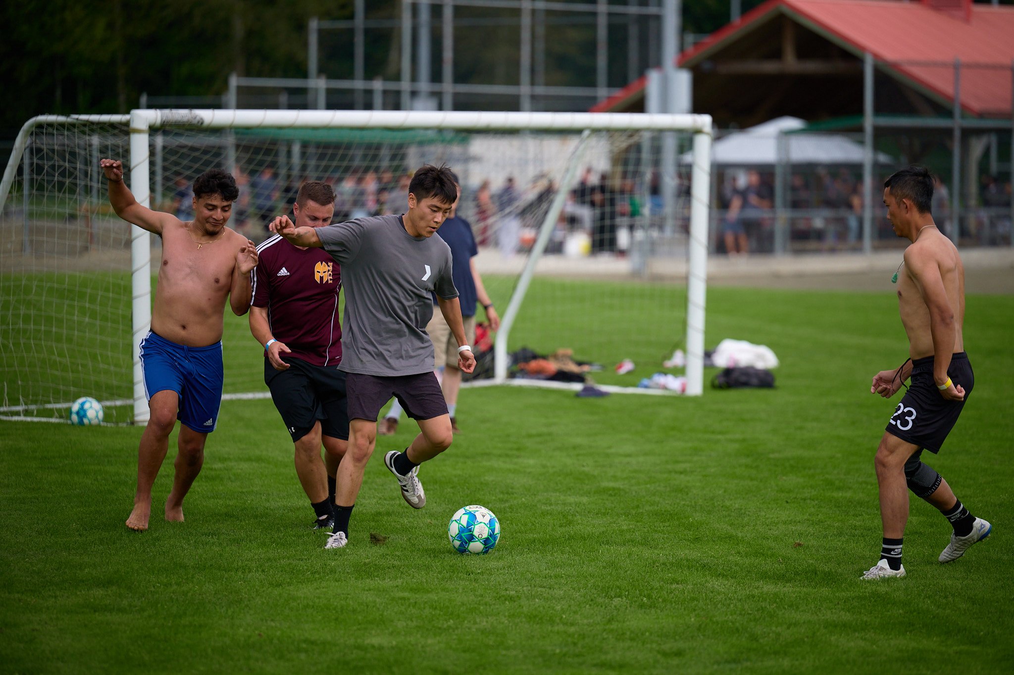 Four men, some shirtless, playing soccer on a grassy field with a goalpost in the background.