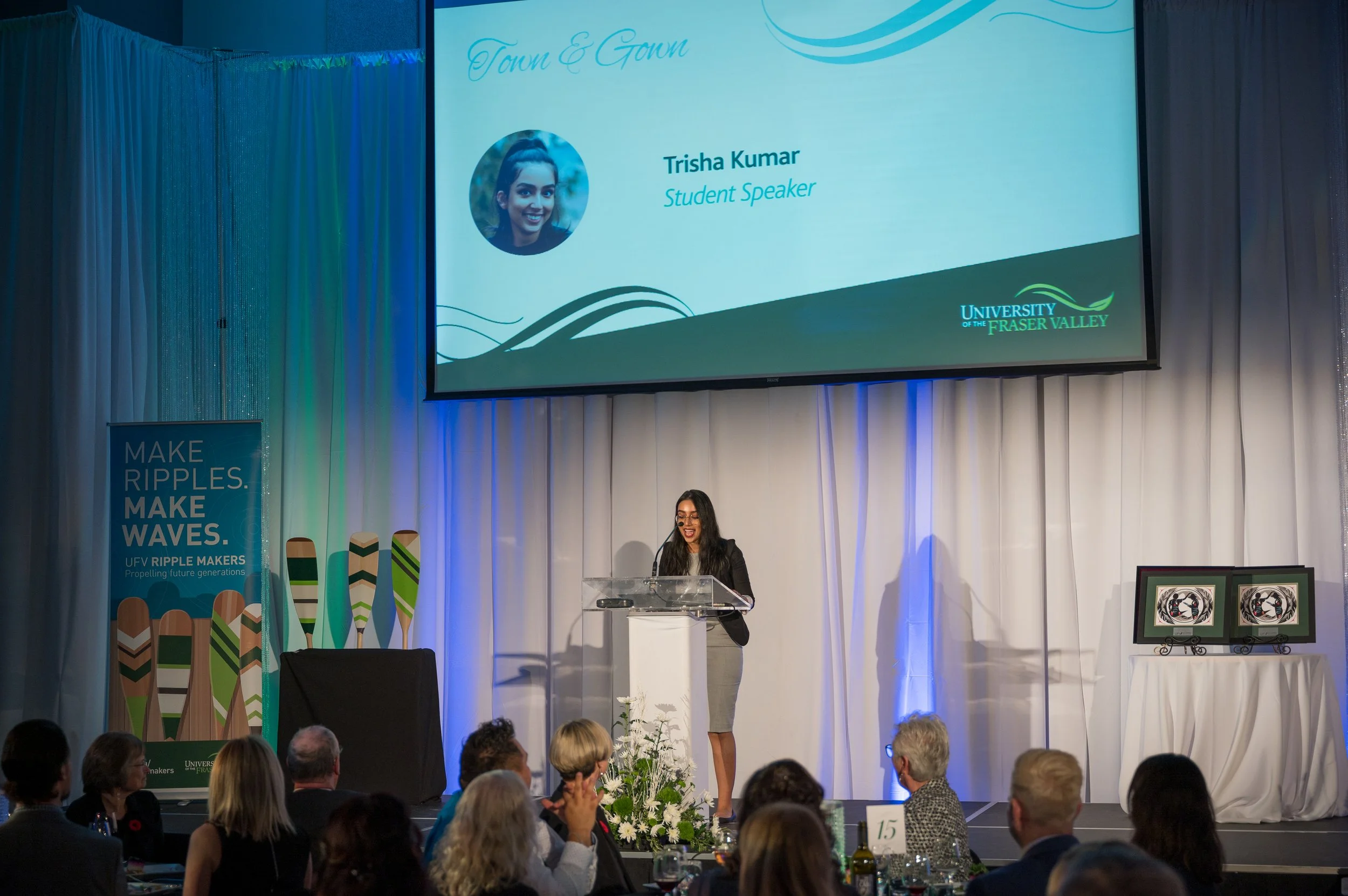 A woman giving a speech at a podium during an event, with an audience seated at tables; a large screen behind her displays her name, photo, and title as a student speaker at the University of the Fraser Valley; a banner to the left promotes UFV rippl