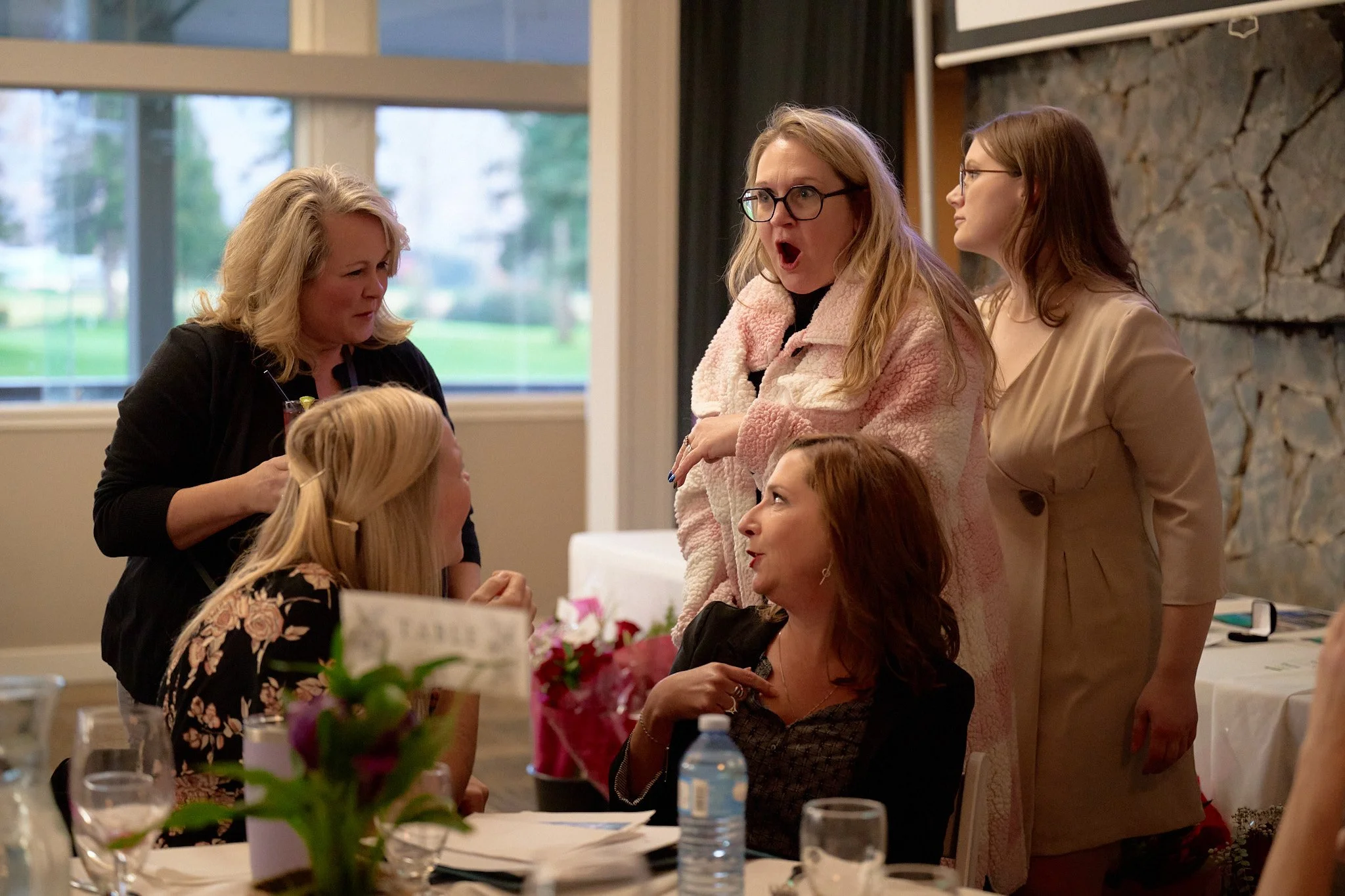 Five women having an animated conversation at a social gathering, with one woman appearing surprised or upset, sitting and standing around a table with flowers and drinks.