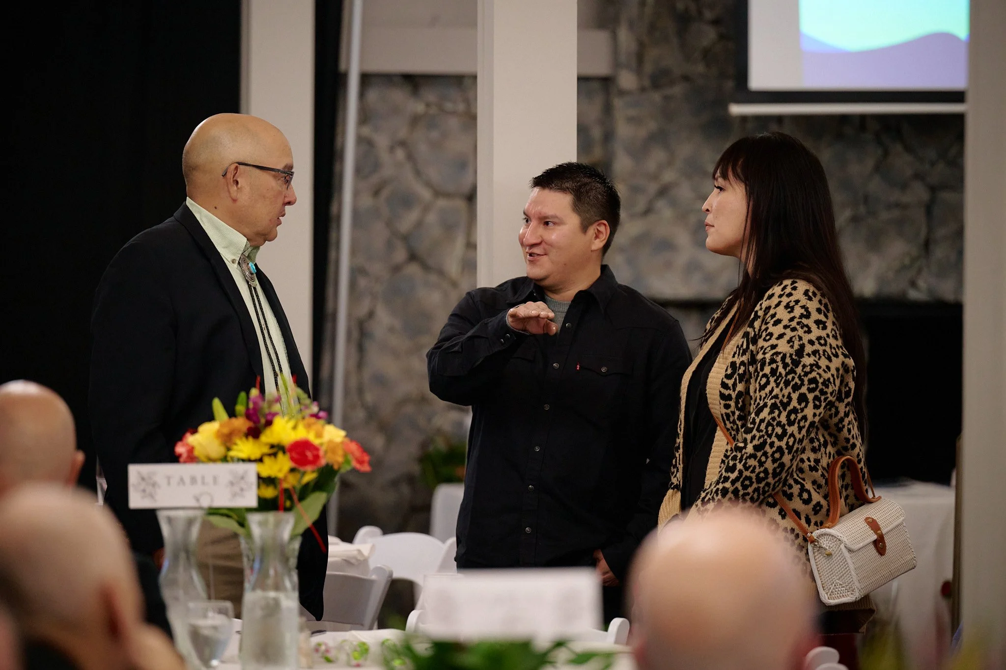 Three people having a conversation at a formal event, with tables and flowers in the foreground.
