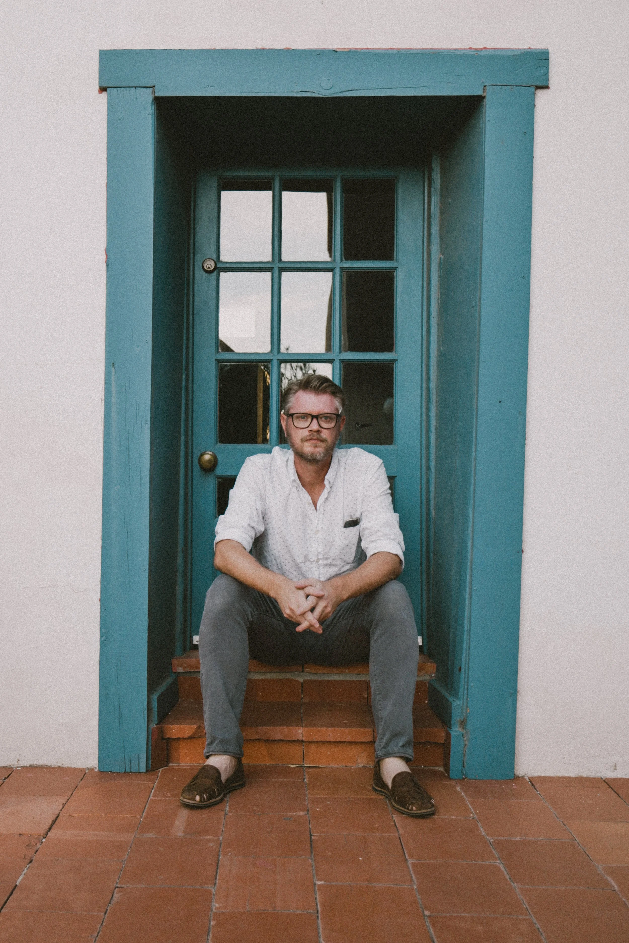 A man with glasses, wearing a white shirt and gray pants, sitting on a small bench with a brick floor, under a blue wooden structure with glass windows, against a white wall.
