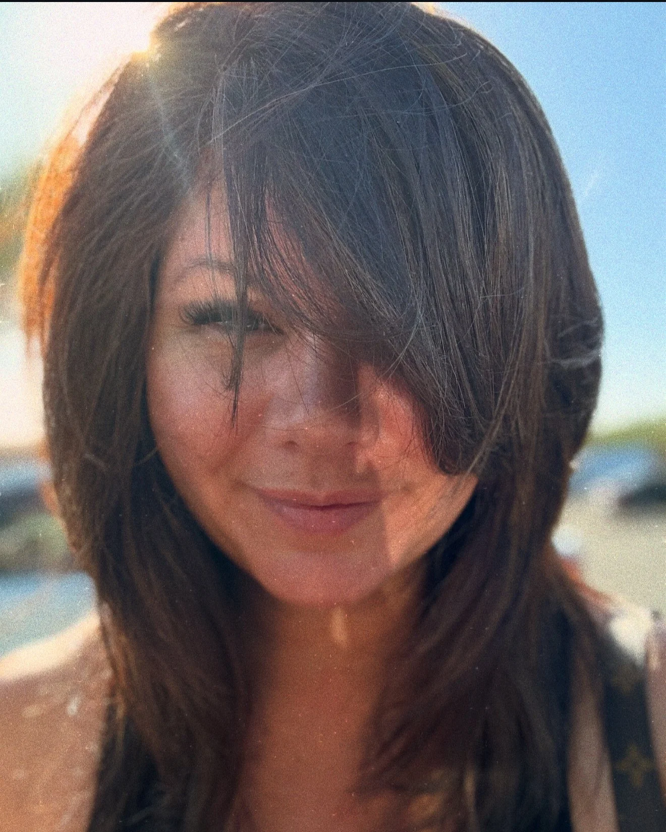 A woman with long dark hair smiling outdoors on a sunny day.