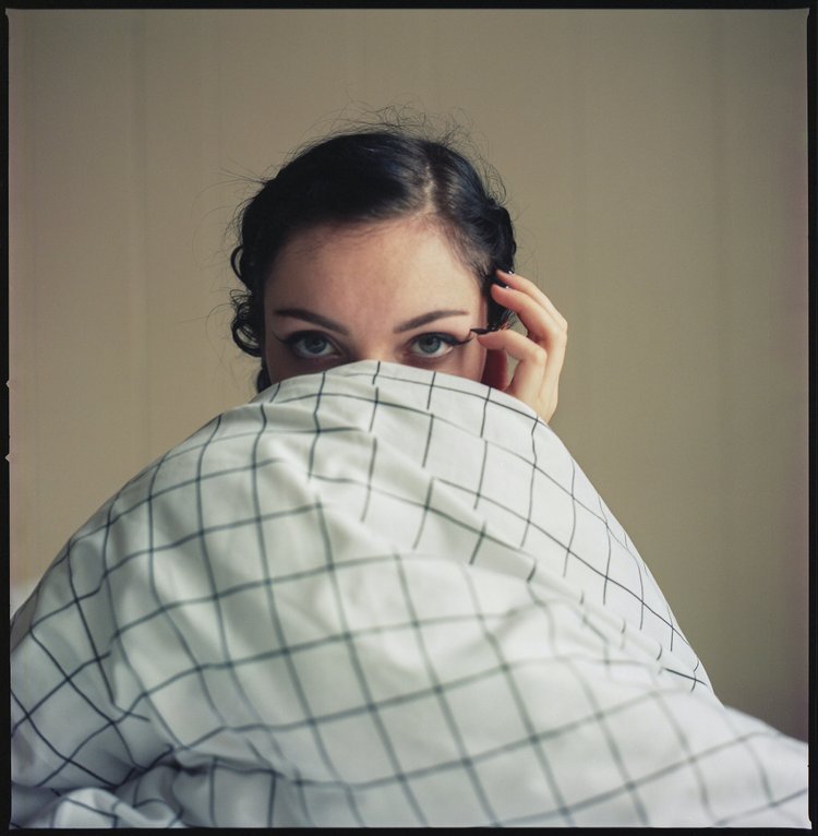 A woman with dark hair peeking over a checkered pillow or blanket, looking directly at the camera with partially covered face.